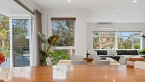 Ein modernes Wohnzimmer mit einem Holzes Esstisch, einer Couch und großen Fenstern mit Blick ins Freie.