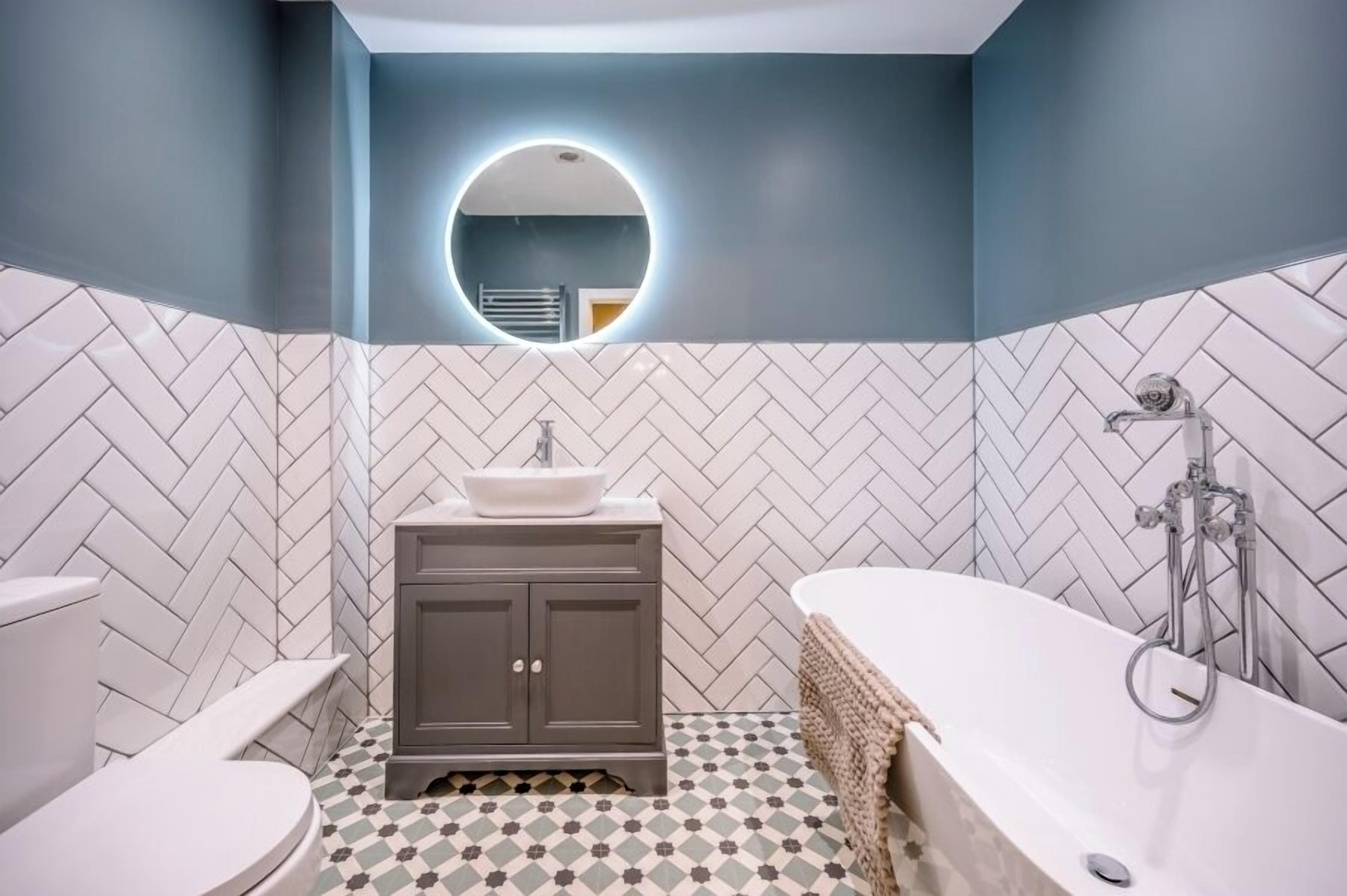 A modern bathroom with a herringbone tile floor, a round mirror, a white sink, a dark cabinet, and a freestanding bathtub.