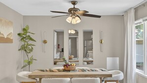 Un comedor con una mesa de madera, sillas blancas acolchadas, un ventilador de techo y un espejo grande que refleja la estancia.