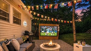 Un patio al aire libre con una pantalla de televisión mostrando un partido de fútbol, rodeado de banderas, un brasero y mobiliario de exterior.