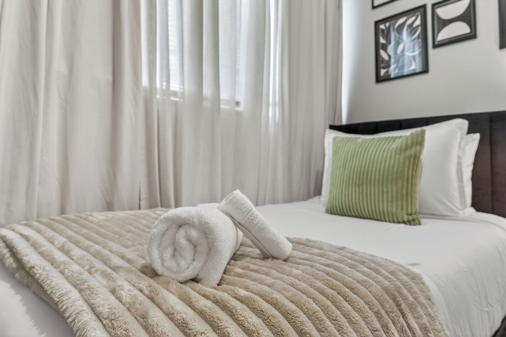 Ein Hotelzimmer mit einem Bett, einem zusammengerollten Handtuch, einem grünen Kopfkissen und einem Fenster mit Vorhängen.