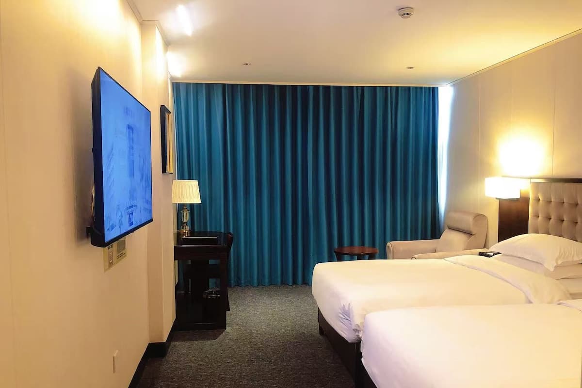 Habitación de hotel con dos camas, televisión, escritorio con lámpara y ventana con cortinas azules.