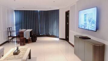 A modern living room with a flat-screen TV mounted on the wall, a dining area with a table and chairs, and a large window with blue curtains.