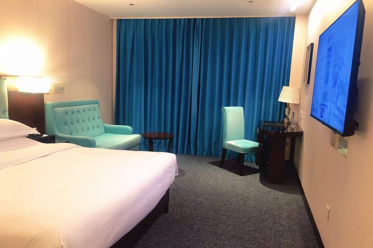 Una habitación de hotel con una cama grande, dos sillones, una mesita, un escritorio con lámpara y un televisor en la pared.