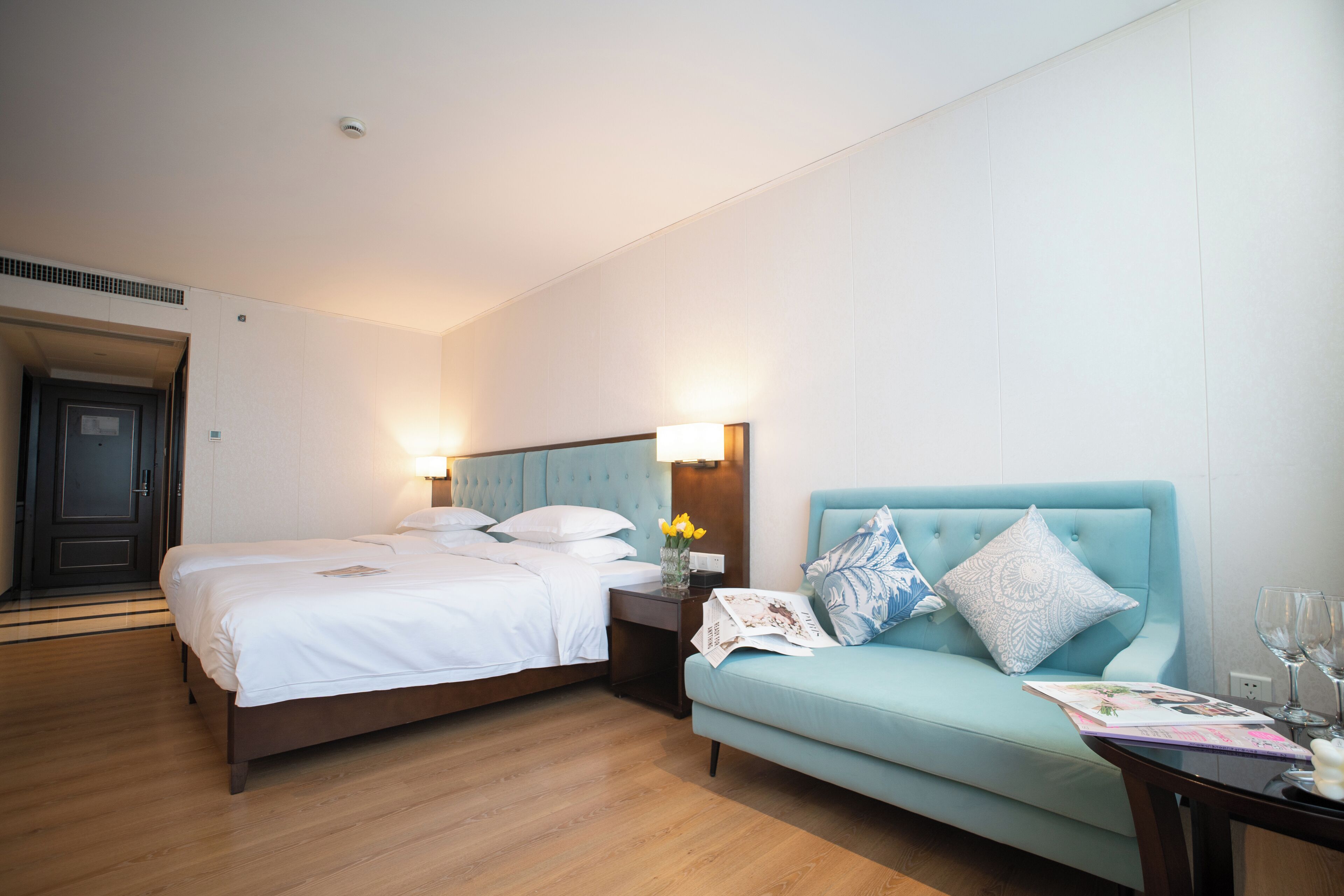 A hotel room with a bed, a sofa, a nightstand, and a table with a magazine and glasses.