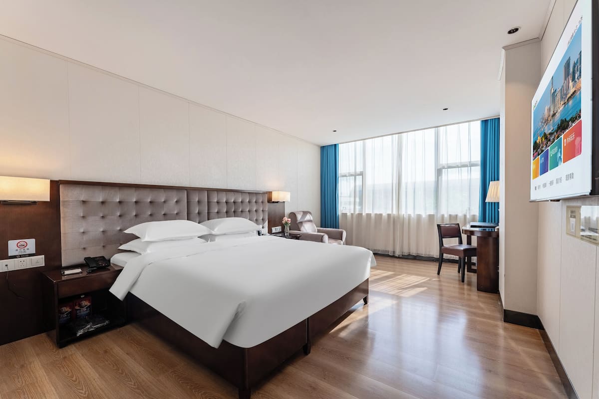 Habitación de hotel con una cama grande, un escritorio y una silla. Hay una ventana con cortinas, un televisor montado en la pared y un cartel en la pared.
