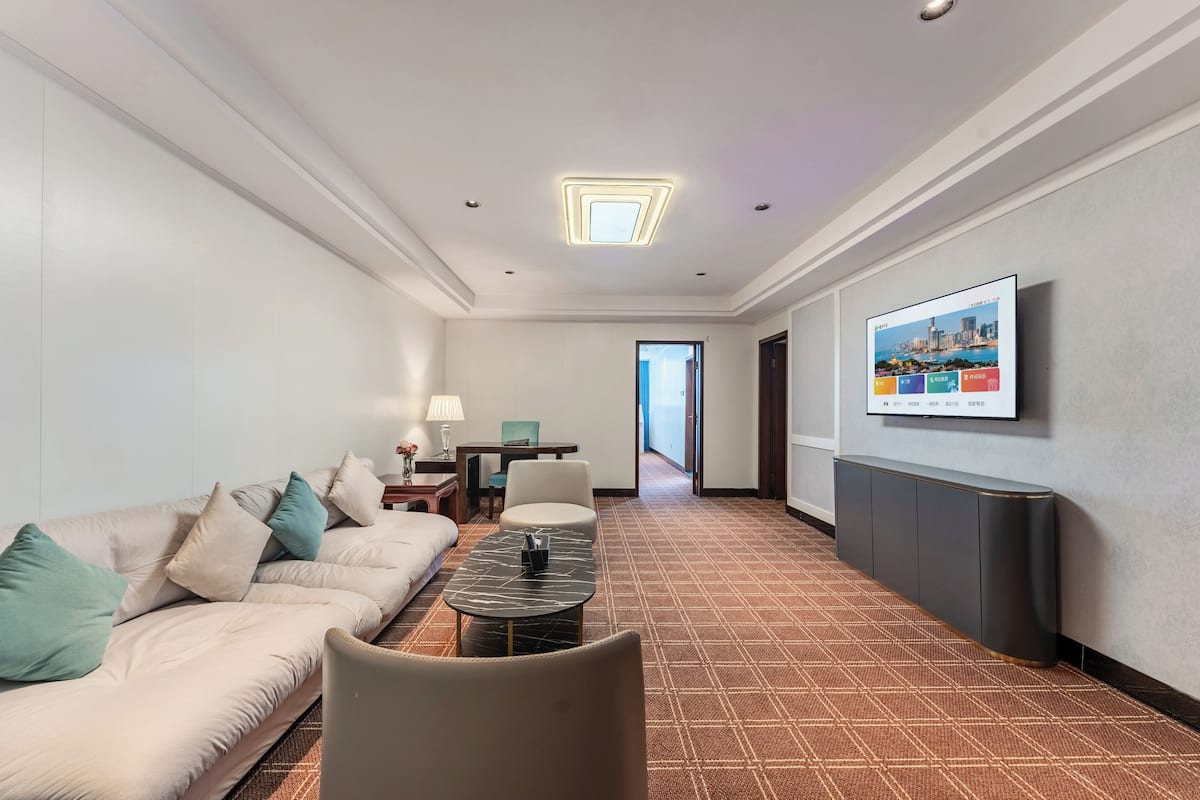 Habitación de hotel con sofá, sillón y una mesita con lámpara. Hay un televisor montado en la pared y una puerta que da a otra habitación.