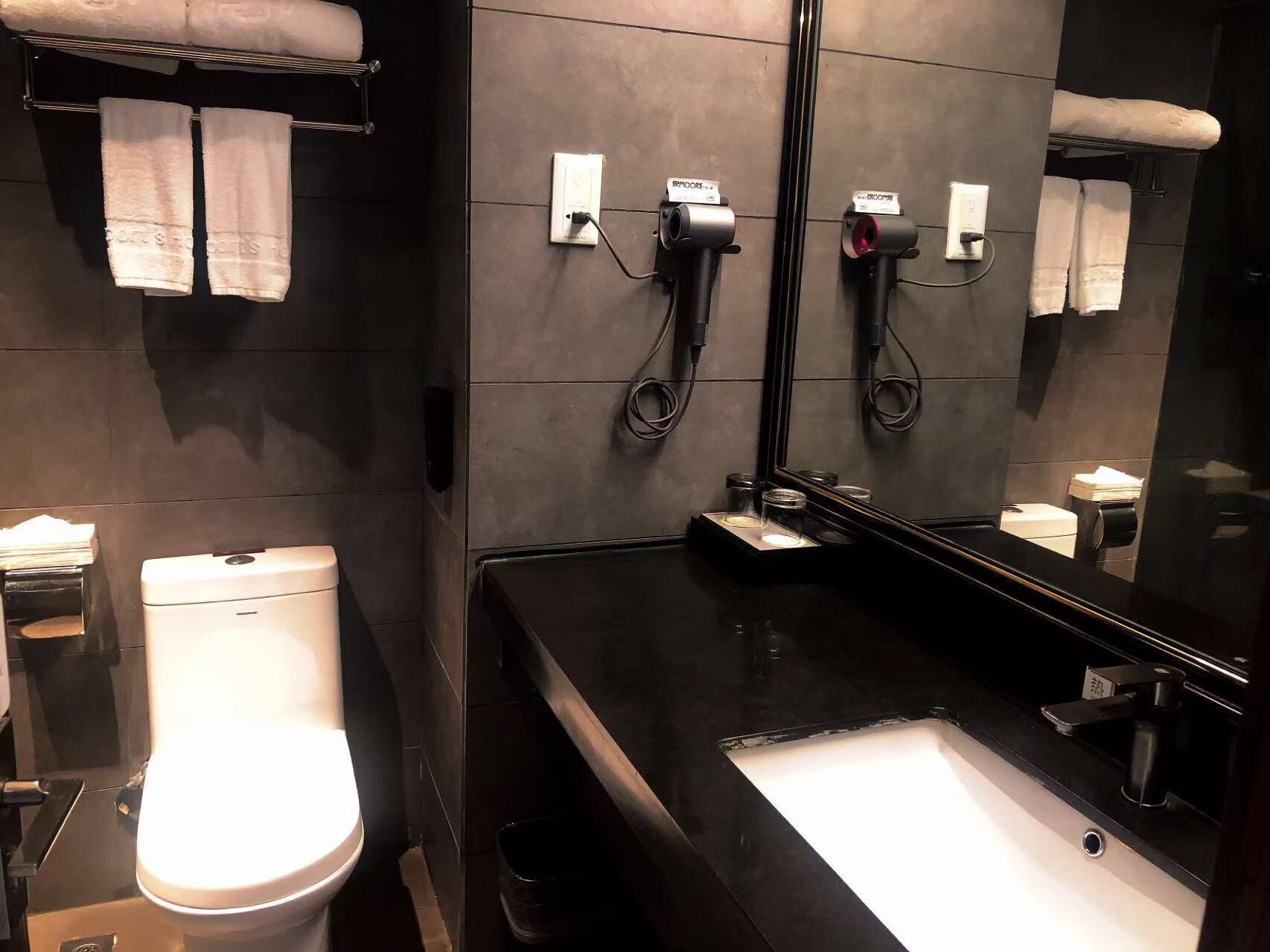 Un baño moderno con un lavabo negro, un inodoro y un espejo. Hay toallas colgadas en un perchero y un secador de pelo en la pared.