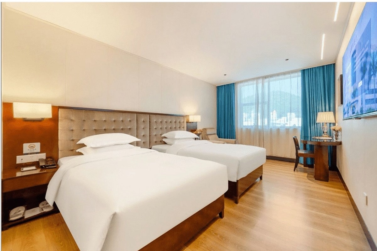 Habitación de hotel con dos camas, cabecero de madera, escritorio con silla y un gran ventanal con cortinas azules.