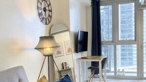 A modern living room with a sofa, a lamp, a flat-screen TV, and a large window with a view of the city. - Modern Central 1BR Apt - Prime City of BGC, Manila (Taguig)