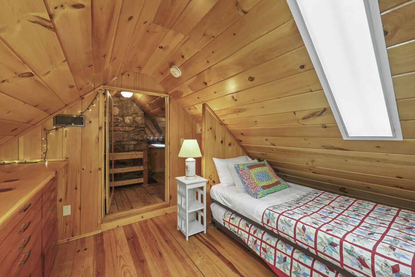 A wooden cabin bedroom with a lofted sleeping area, a small bedside table with a lamp, and a quilted bedspread.