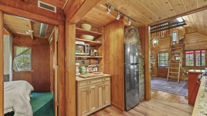 A wooden cabin interior with a kitchen area, a bed, and a view of the outside.