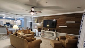 Un salon moderne avec un grand canapé, une table basse et un téléviseur fixé à un mur en bois. On aperçoit un ventilateur de plafond et un coin repas à l’arrière-plan.