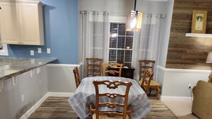 Un espace repas avec un plancher en bois, une table entourée de chaises, un coin cuisine avec des placards et une fenêtre agrémentée de rideaux.