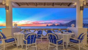 Un espace repas avec des chaises et des tables blanches, offrant une vue panoramique sur la côte et un magnifique coucher de soleil.