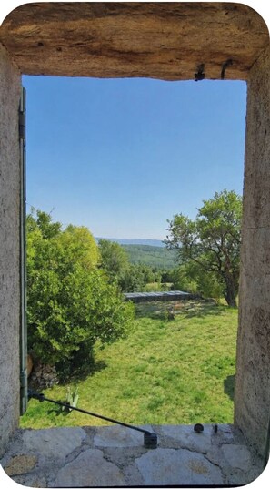 Une fenêtre encadrée de pierres donnant sur un paysage verdoyant, avec des arbres et une chaîne de montagnes lointaine.