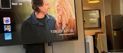 A hotel room with a large flat-screen TV mounted on the wall displaying a movie titled "Virgin River."