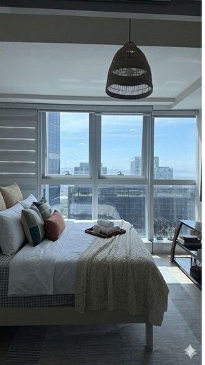 A modern bedroom with a large bed, a hanging light fixture, and a city view through large windows. - Modern Uptown Parksuites - BGC (Taguig)