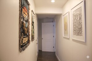 A hallway with framed artwork on the walls, a white door at the end, and a wooden floor. - 3BR Walk-out Retreat-Quiet Country Hills Location (Calgary)