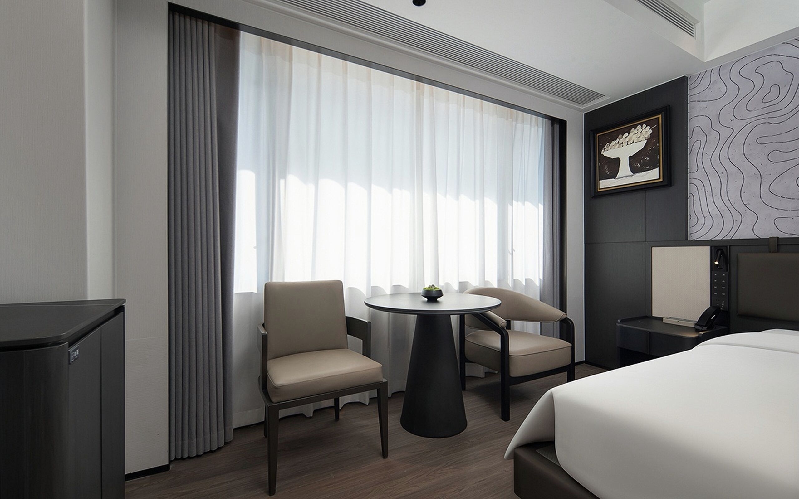 Een moderne hotelkamer met een bed, een ronde tafel met stoelen, een bureau met een stoel en een groot raam met gordijnen.