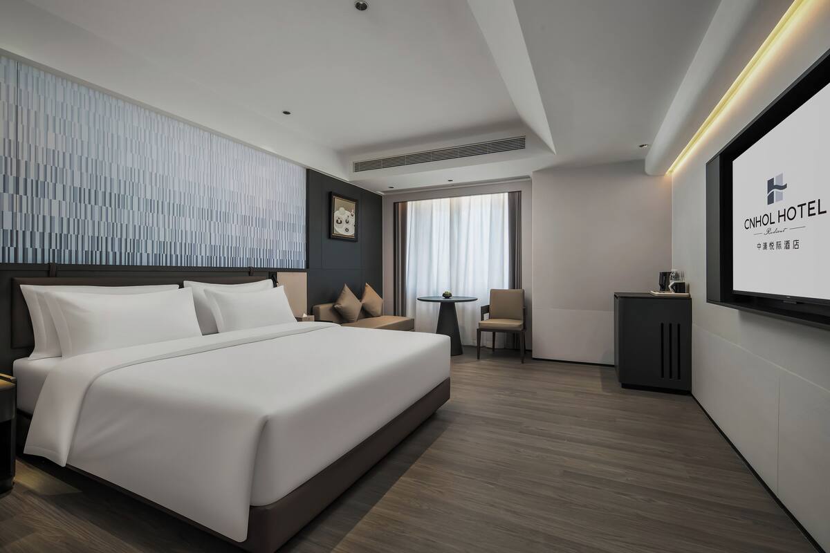 Une chambre d’hôtel moderne, dotée d’un grand lit, d’un bureau, d’une chaise et d’un écran de télévision affichant le nom de l’hôtel.