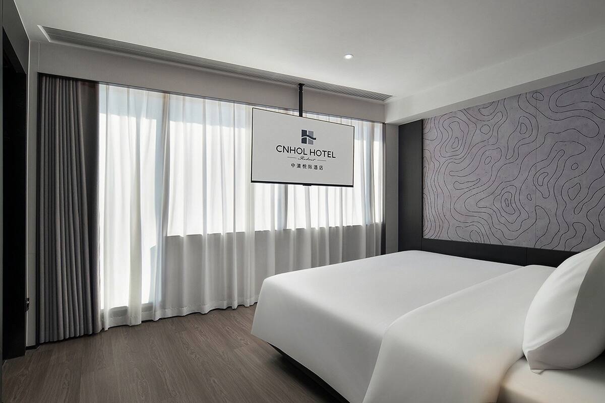 Une chambre d’hôtel moderne dotée d’un grand lit, d’un téléviseur à écran plat fixé au mur et offrant une vue sur la ville grâce à de grandes fenêtres du sol au plafond.