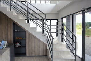 Una escalera moderna con barandillas metálicas, un estante de madera con objetos, y una vista a través de un ventanal.
