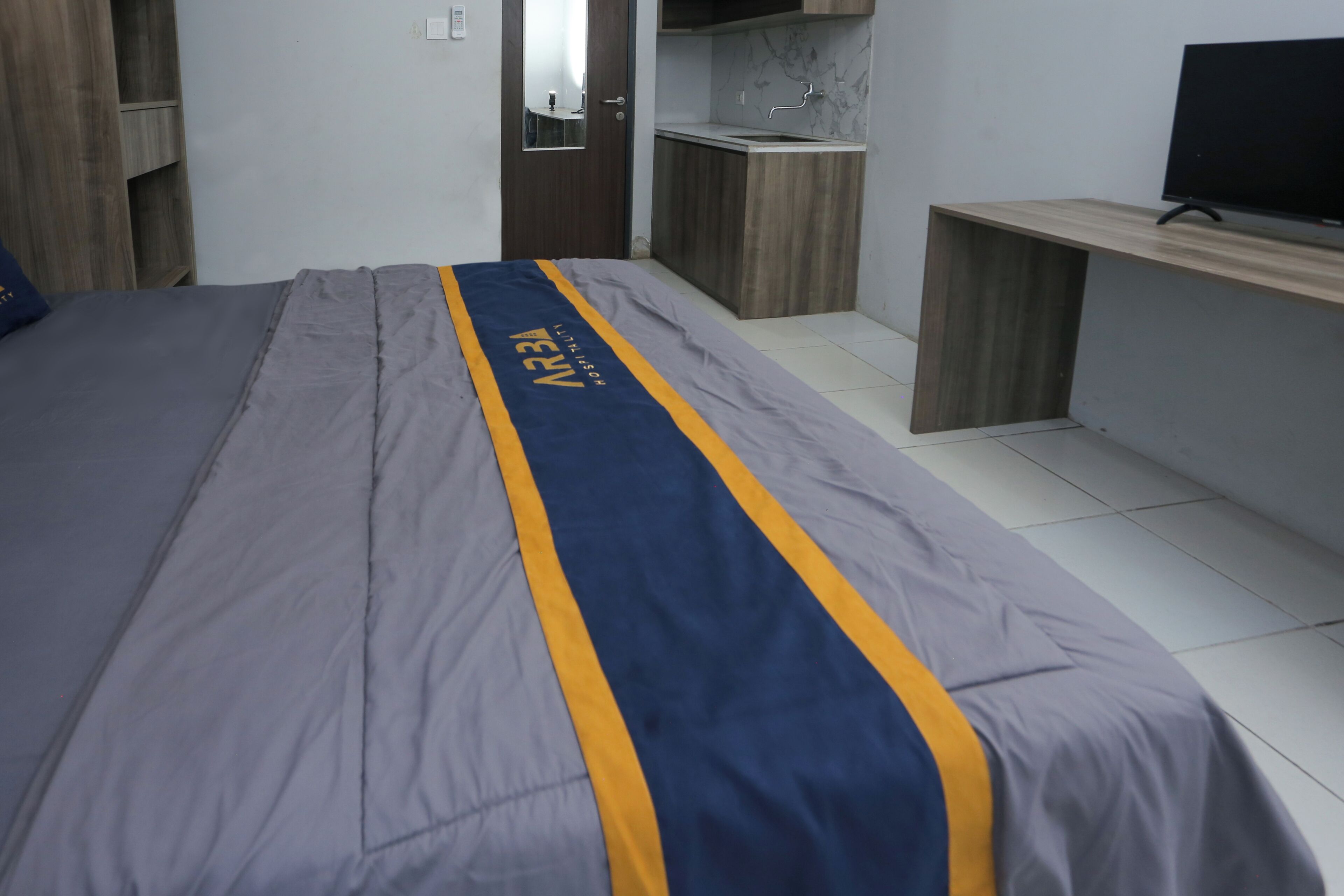 A bedroom with a bed, a TV on a stand, a wooden wardrobe, and a small kitchenette.