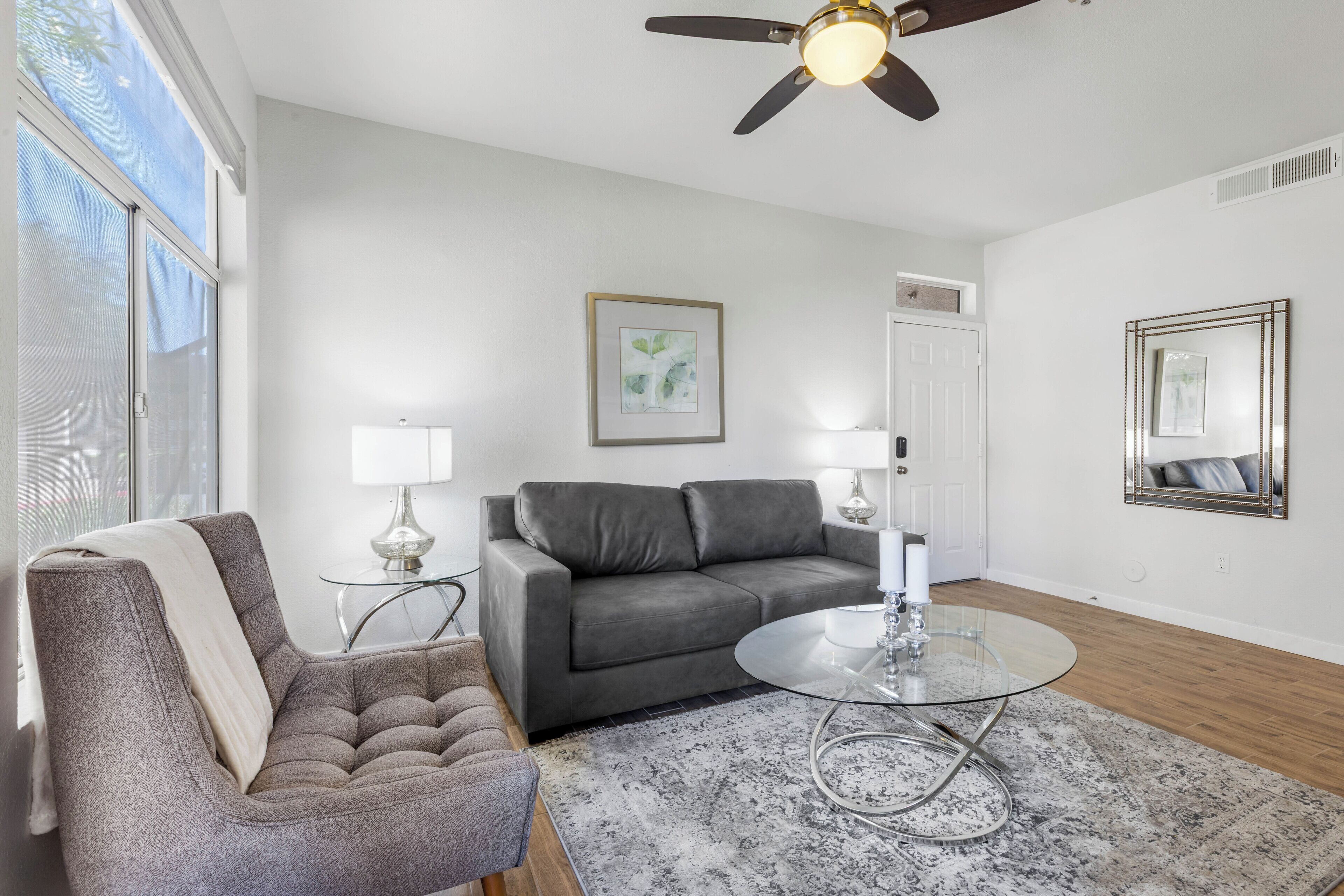 Ein Wohnzimmer mit grauem Sofa, einem runden Glastisch, einem gemusterten Teppich in Grau, einem Deckenventilator und einem gerahmten Bild an der Wand.