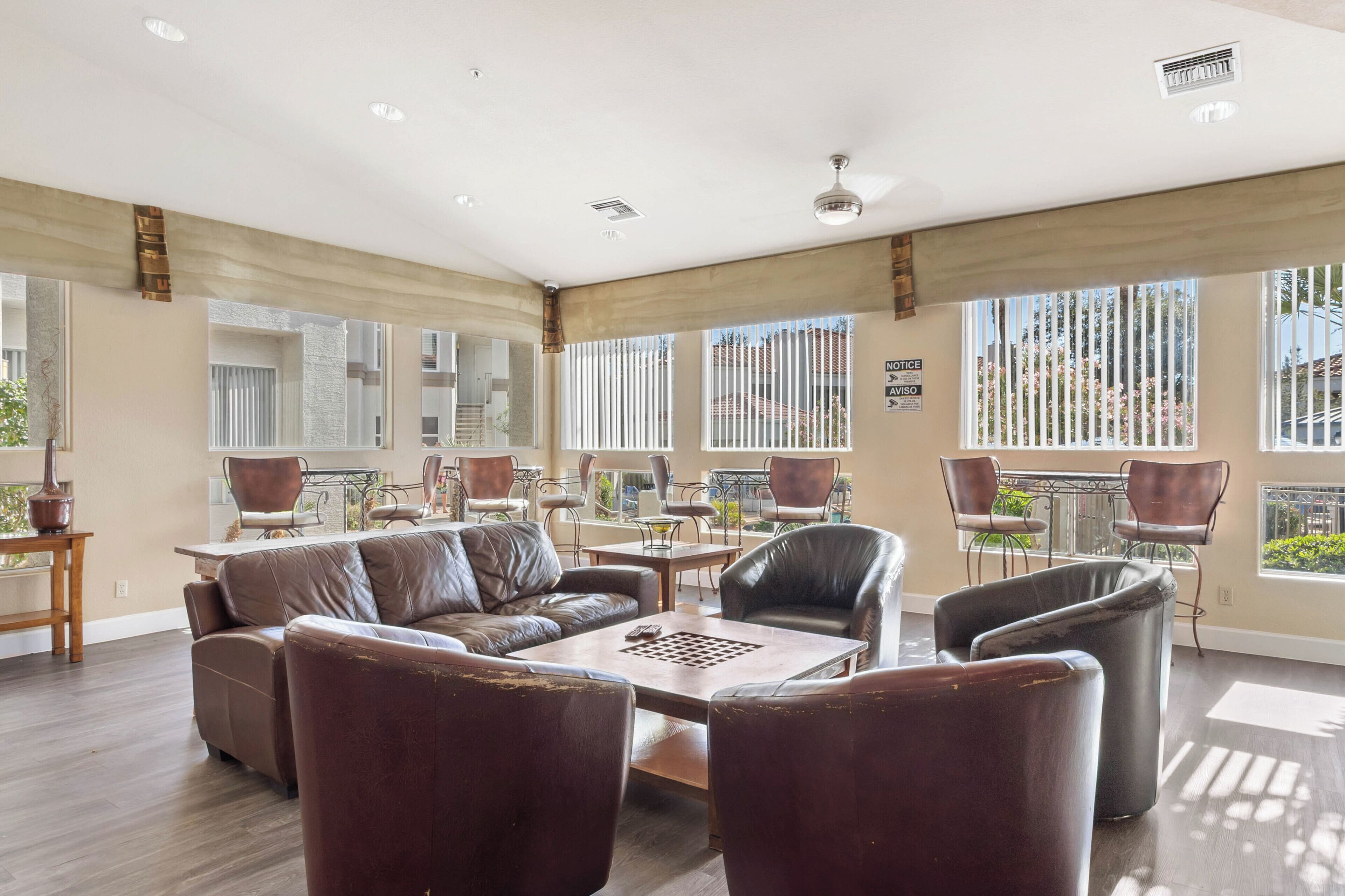 A spacious living area with a leather sofa, a coffee table, and multiple chairs. There are large windows with blinds, a dining area with a table and chairs, and a view of the outside.