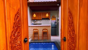 A pool with blue tiles, wooden doors with carvings, a kitchen with wooden cabinets and a refrigerator, and two wooden stools. - Desta House Ubud (Ubud)