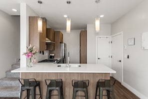 Una cocina moderna con una isla central, taburetes de bar, armarios de madera, electrodomésticos de acero inoxidable, iluminación colgante y una escalera que lleva a otro nivel.