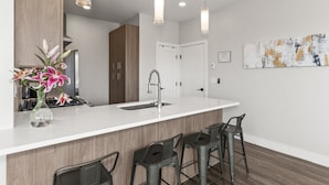 Una cocina moderna con encimera blanca, pileta de acero inoxidable y tres taburetes de bar. Hay un florero con flores sobre la encimera, y lámparas colgantes iluminan el espacio.