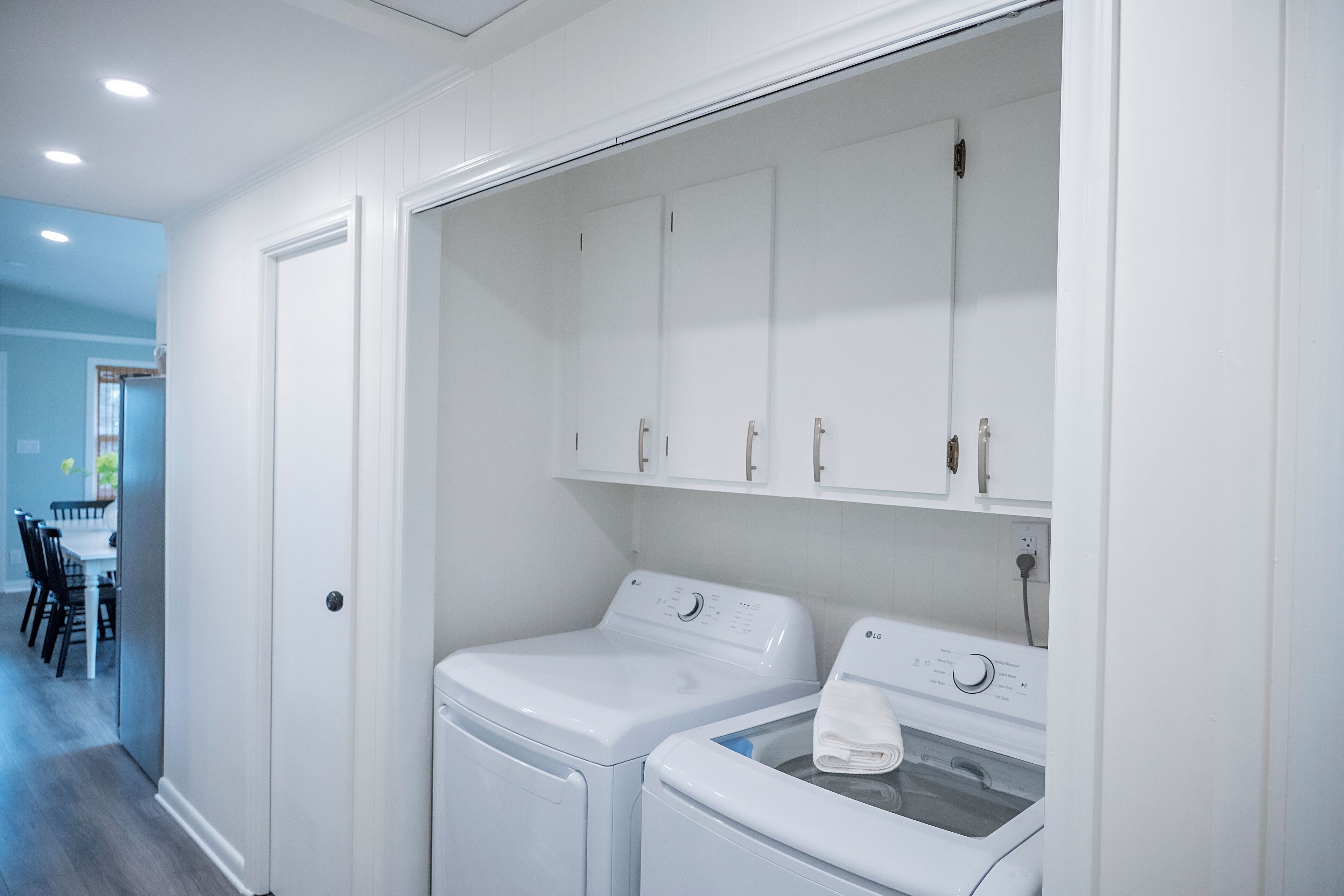 A laundry room with two front-loading washing machines and a dryer, white cabinets above, and a door leading to a dining area.