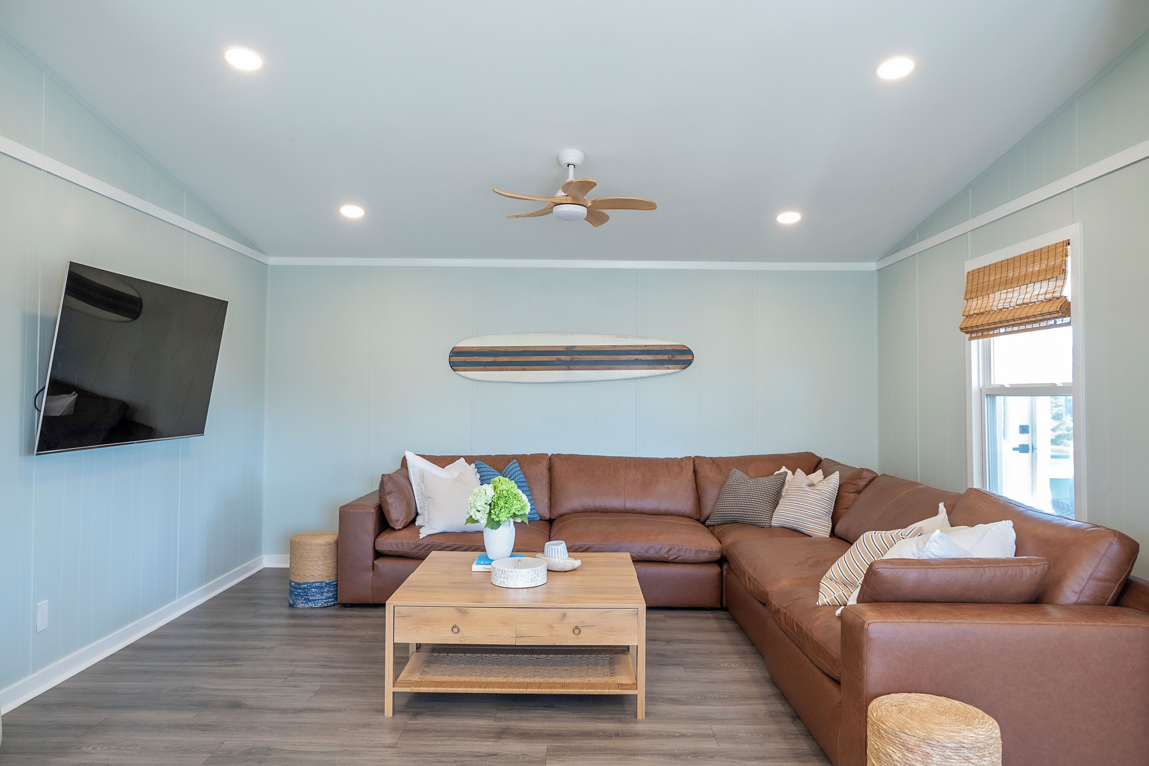 A living room with a brown leather sofa, a wooden coffee table, a flat-screen TV mounted on the wall, and a ceiling fan.
