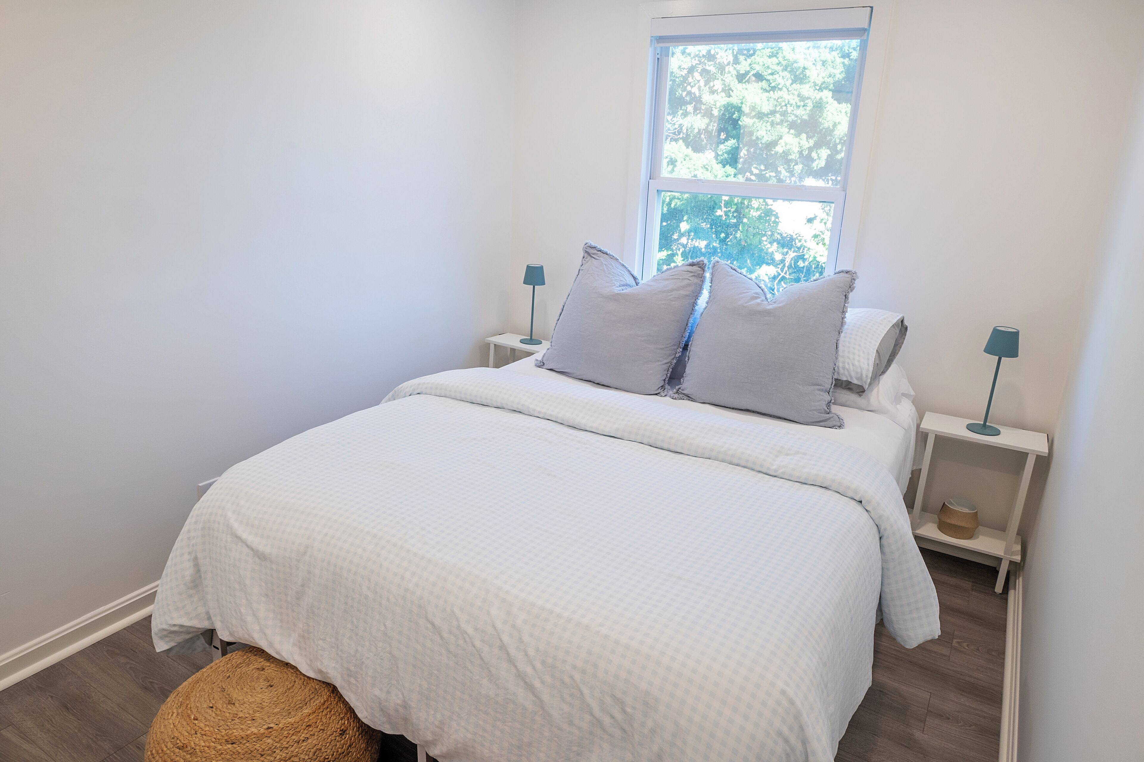 A neatly made bed with a white bedspread and two pillows, flanked by nightstands with lamps, in a room with a large window and a woven basket on the floor.