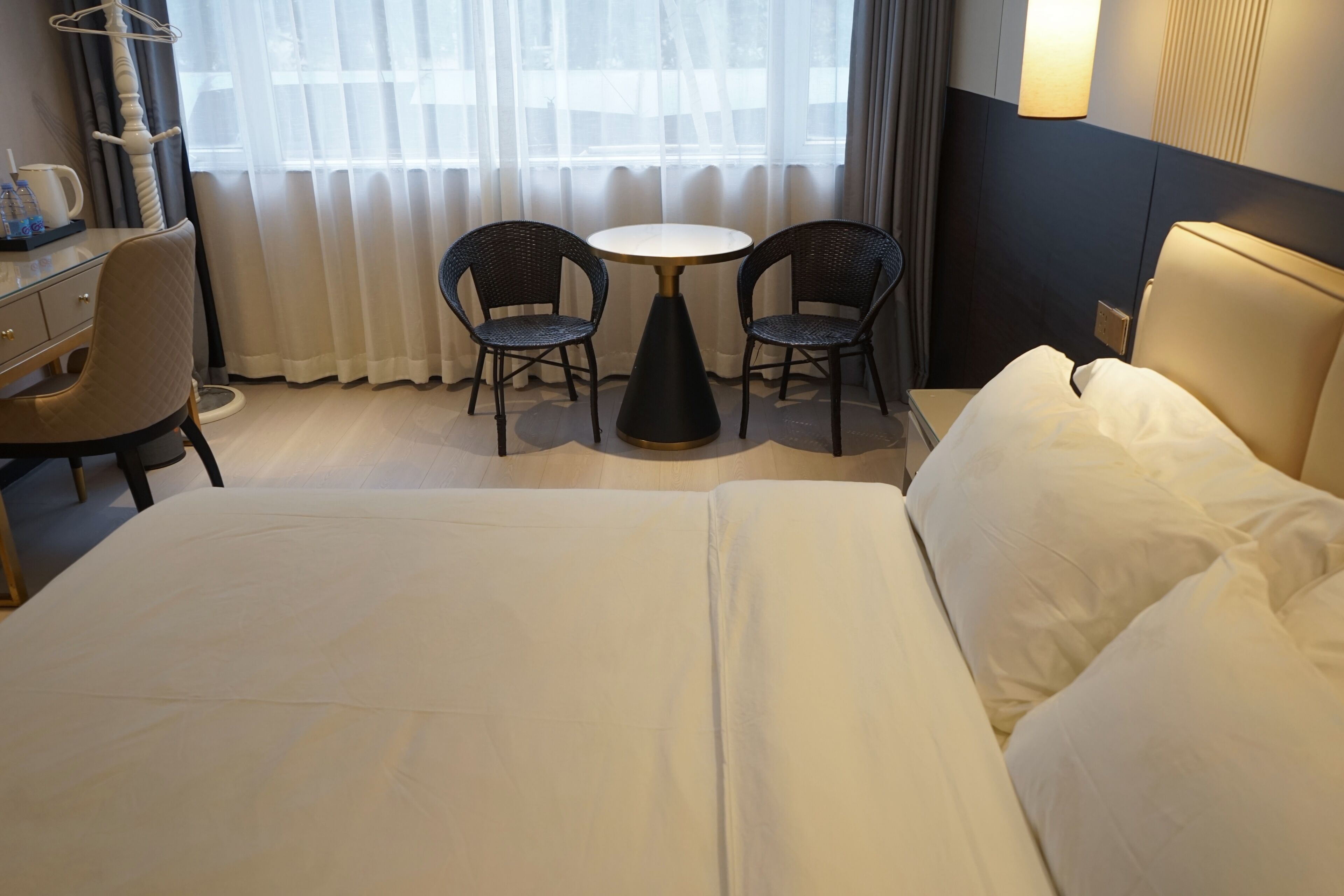 A hotel room with a bed, two chairs around a small table, a desk with a chair, and a window with curtains.