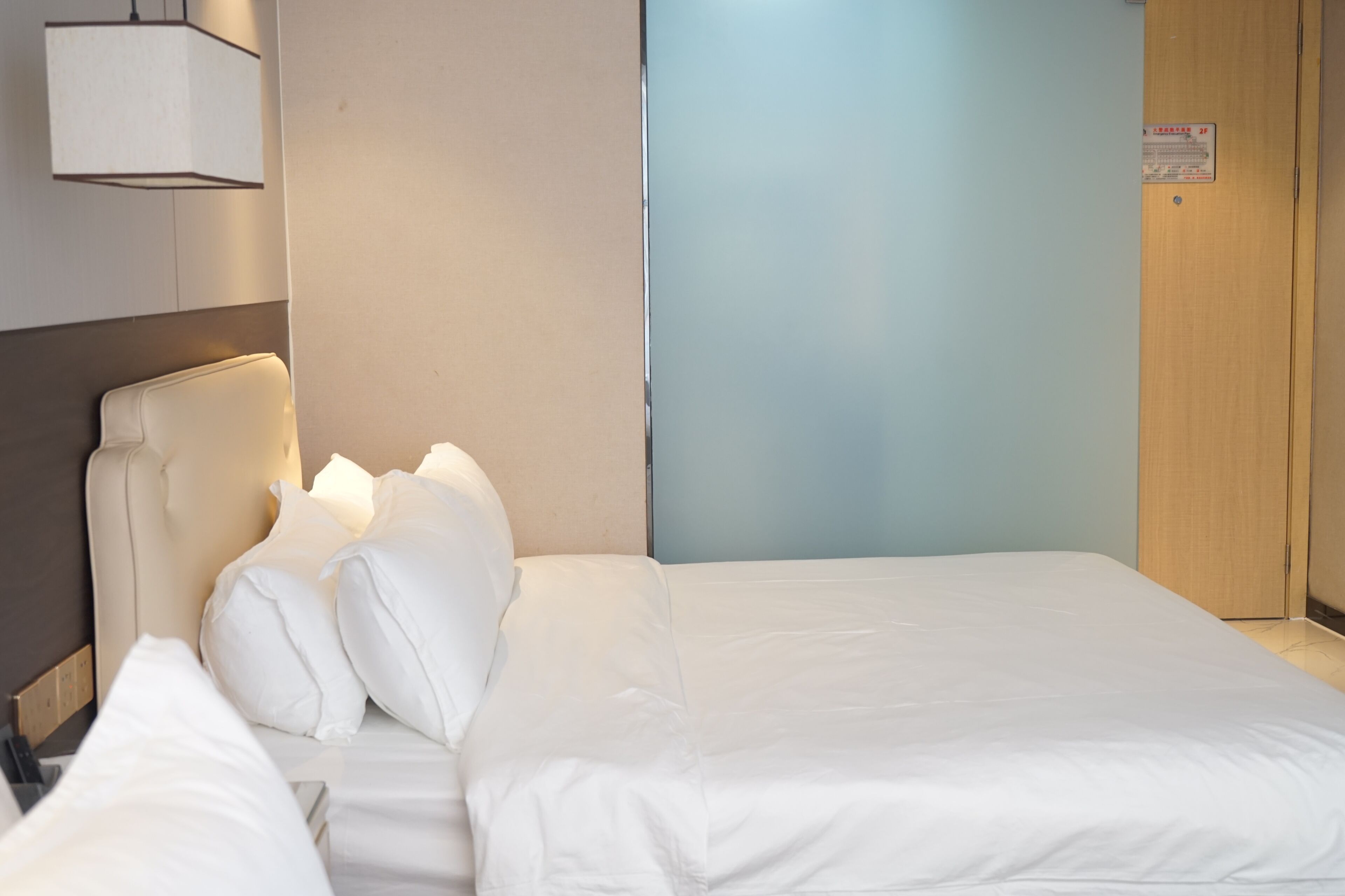 A hotel room with a bed, a headboard, a wall-mounted lamp, and a door.