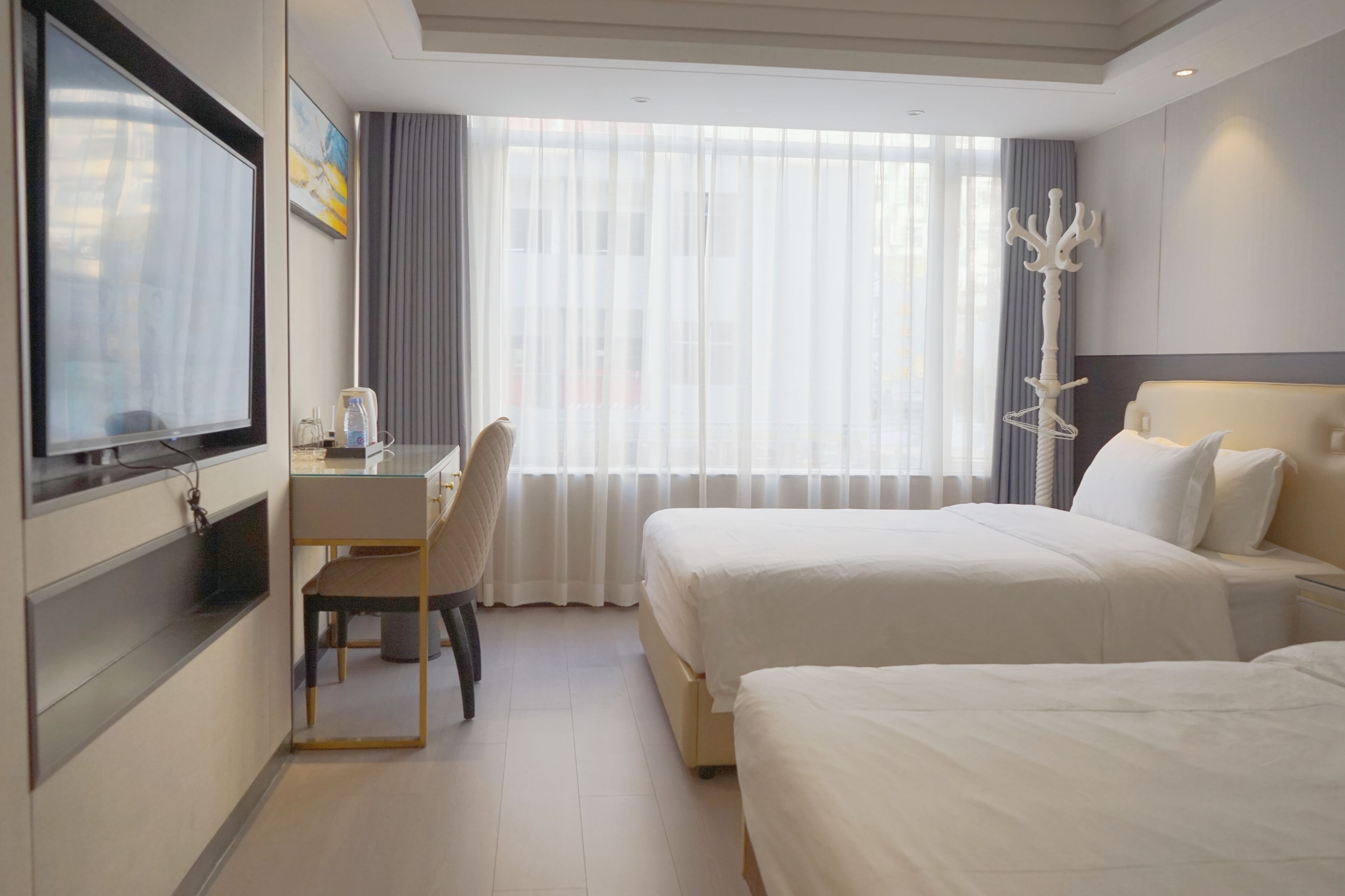 A hotel room with two beds, a desk, a chair, a TV, and a large window with curtains.
