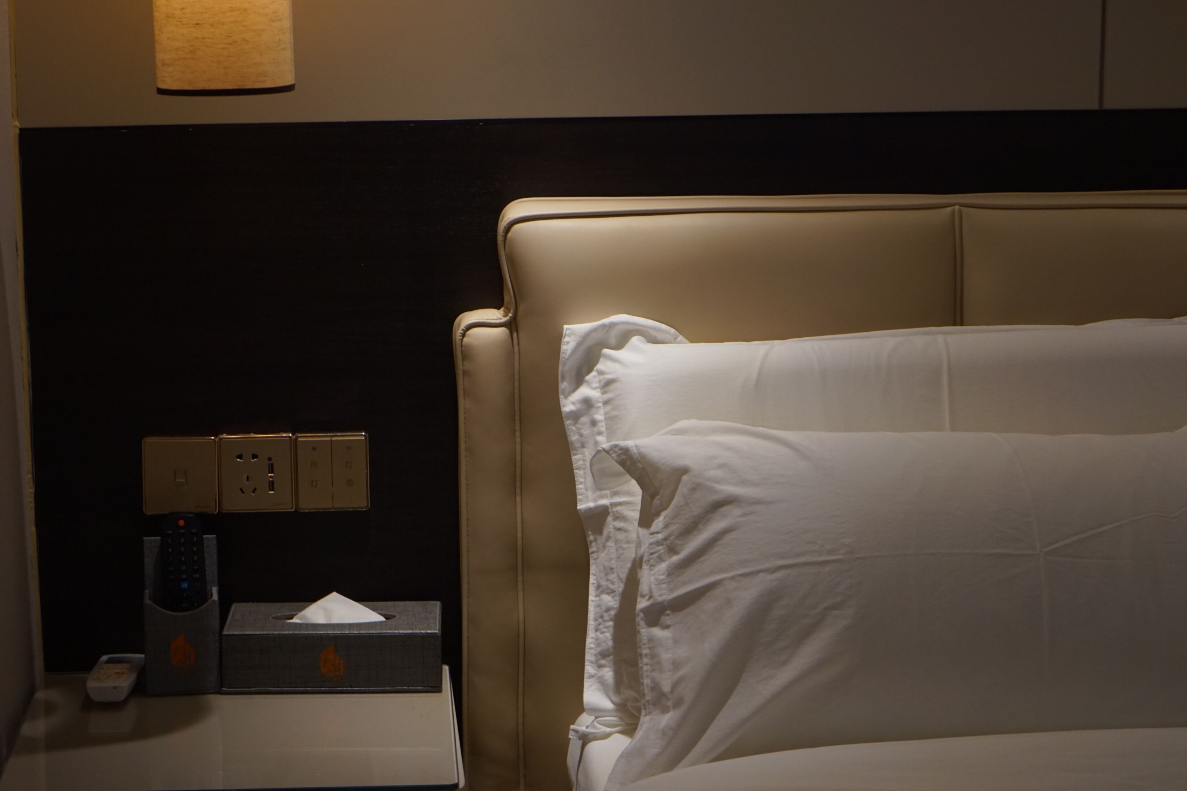 A hotel room with a bed, a nightstand with a remote control, a tissue box, and a wall socket.