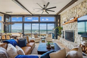 A spacious living room with a large window overlooking the ocean, a stone fireplace, and a ceiling fan. - Pacific Paradise - Arch Cape Beachfront Home with Fireplace! (Arch Cape)