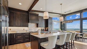 Una cucina moderna con pensili in legno scuro, una grande isola con sgabelli da bar e vista sull'oceano attraverso ampie finestre.