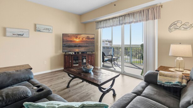A living room with a sofa, a coffee table, a TV, and a balcony with a view. - Welcome to Oceanwalk 15-505! (New Smyrna Beach)