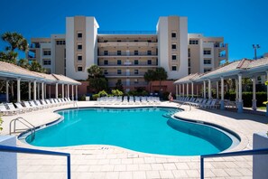 A swimming pool with lounge chairs, a covered area, and a multi-story building in the background. - Welcome to Oceanwalk 15-505! (New Smyrna Beach)