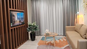 Ein Wohnzimmer mit an einer Holzverkleidung montiertem Fernseher, einem Topf mit Pflanze, einem runden Tisch mit Fernbedienung, einem Sofa und einem großen Fenster mit Vorhängen.