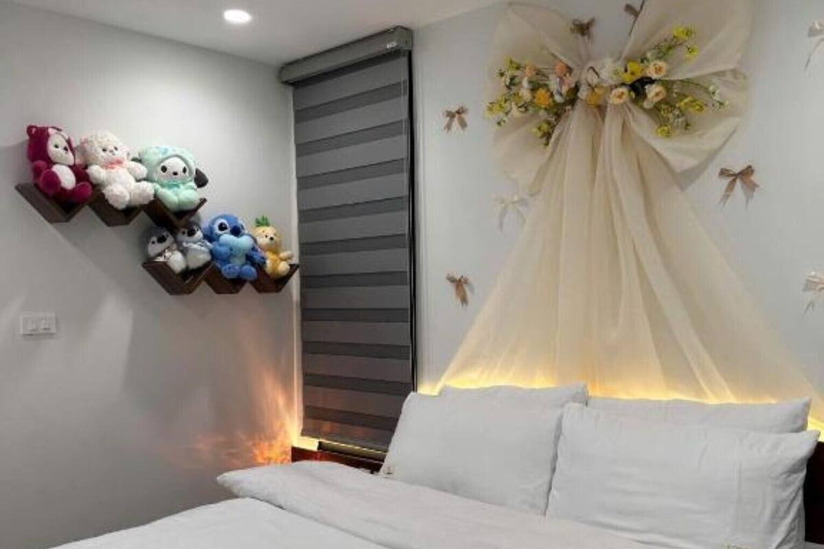 Un dormitorio con una cama, un estante con juguetes de peluche, una ventana con persianas y un arreglo floral decorativo.