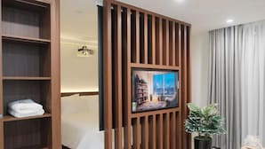 Ein Hotelzimmer mit Bett, einer Holzabtrennung, einem Fernseher, einer Zimmerpflanze und einem Fenster mit Vorhängen.
