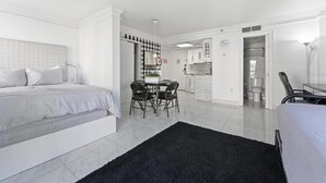 A modern, well-lit room with a bed, a dining table with chairs, and a kitchen area.