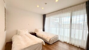 Ein Hotelzimmer mit zwei Betten, Holzboden, einem großen Fenster mit durchscheinenden Vorhängen und einer Decke mit Einbautenleuchten.