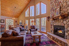 A spacious living room with a stone fireplace, large windows, and a high ceiling. - Alpine Escape | 3BR/3BA Cabin Near Breckenridge (Fairplay)